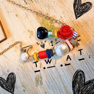 May include: Two gold-coloured necklaces with beaded pendants. One pendant has a black heart-shaped bead, a red rose, and various colourful beads. The other pendant has a pearl and the letters T.W.A.T. in Morse code.