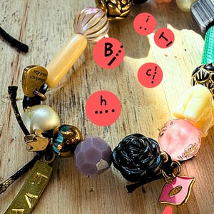 Op de afbeelding: Een kralenarmband met diverse kleurrijke kralen, waaronder goud, zwart, roze en paars. De armband heeft een zwarte rozenkraal, een gouden bedel met het woord "FAB" en een roze lippenbedel. De kralen zijn geregen op een zwart koord.