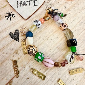 May include: A colourful beaded bracelet featuring pink, green, gold, and black beads. The bracelet includes charms inscribed with "HOT", "CUTE", "SEXY", and "FAB". A wooden heart with the word "HATE" is in the background.