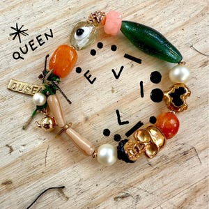 May include: A colourful beaded bracelet with orange, green, and pearl-like beads. The bracelet features a gold-coloured skull charm and a small gold-coloured teapot charm. The word "QUEEN" is written on the bracelet.