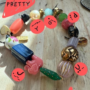 Op de afbeelding: Een kleurrijke kralenarmband met een verscheidenheid aan vormen en kleuren. De armband bevat roze, blauwe, groene, gouden en parelachtige kralen. Het woord "PRETTY" staat in het rood op de afbeelding.