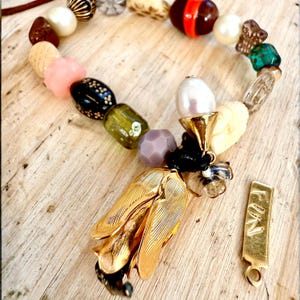 May include: A distinctive necklace with a mix of beads in cream, pink, black, green, and clear. It features a gold-tone leaf pendant and a small gold tag with the word "FUN".