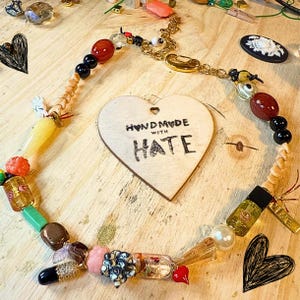 Può includere: Una collana fatta a mano con una varietà di perline rosse, nere, dorate e trasparenti. La collana è disposta a forma di cuore attorno a un cuore di legno con la scritta "HANDMADE WITH HATE".