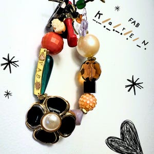 May include: A vibrant charm bracelet featuring a black flower pendant with a pearl, various beads in orange, gold, and a green and gold charm with "FAB". The word "FAB" is also written in Morse code.