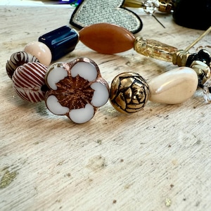 May include: A close-up of a beaded necklace with various beads in shades of brown, white, gold, and navy blue. The necklace features a flower-shaped bead with white petals and a copper centre, along with other unique, decorative beads.