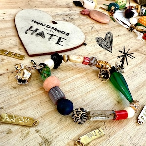 May include: A vibrant beaded bracelet featuring a mix of shapes and colours, including green, pink, and gold beads. A wooden heart with the words "HANDMADE WITH HATE" is in the background, alongside small gold charms and other beads. The bracelet rests on a wooden surface.