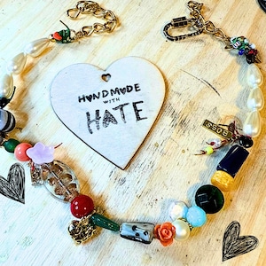 Può includere: Una collana fatta a mano con una varietà di perline, tra cui perle, perline di vetro colorate e una catena color oro. Un'etichetta a forma di cuore in legno con la scritta "HANDMADE WITH HATE" è al centro. La collana ha una chiusura color oro.