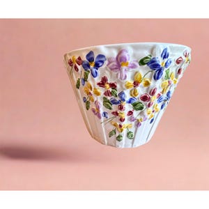 Peut inclure: Un bol en céramique blanche doté d'une surface extérieure cannelée, orné de motifs floraux en relief peints à la main dans des tons de bleu, jaune, violet et rouge foncé, agrémentés de feuilles vertes pour un design botanique texturé.