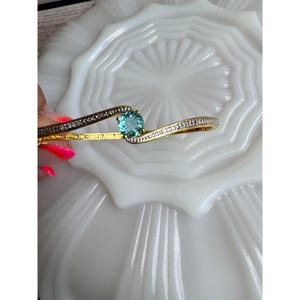 May include: A gold-tone hinged bangle bracelet featuring a central round-cut teal gemstone. The bracelet has a curved, bypass-style design with pavé-set clear stones lining the top edges of the band, creating a shimmering, elegant appearance.