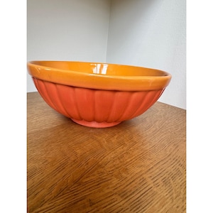 May include: A large, orange and yellow ceramic mixing bowl. The bowl has a ribbed exterior and a smooth, glossy interior. The rim is a bright yellow, while the body is a vibrant orange. The bowl is sitting on a wooden surface.