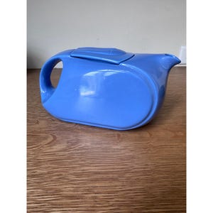 May include: A smooth, cornflower blue ceramic teapot with a horizontal, elongated oval body. It features a curved handle integrated into the side, a matching lid with a rectangular top detail, and a short, pointed spout on the right side.