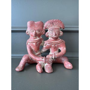 May include: A terracotta-colored ceramic sculpture depicting two seated figures joined together. The figures feature stylized facial features, necklaces, and one wears a prominent headdress. The surface has a matte, earthy texture with visible wear.