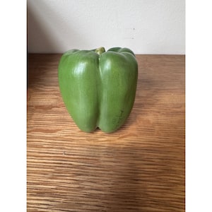 May include: A green bell pepper figurine with a smooth, glossy finish and a small stem at the top. The surface features subtle vertical ridges and a hand-painted signature in black ink on the lower right side, adding detail to the vegetable shape.