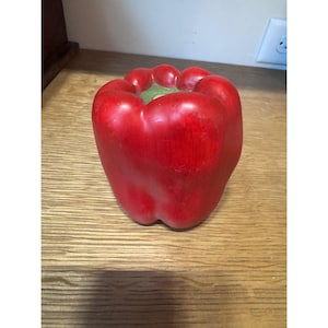 May include: A bright red bell pepper shaped object with a smooth, glossy finish. The top features a recessed green stem area with rounded lobes. The exterior surface shows subtle variations in color intensity, mimicking the appearance of a real vegetable.