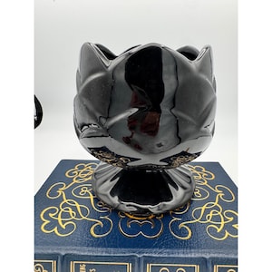 May include: A glossy black ceramic vase with a unique floral design. The vase has a pedestal base and a wide, open top. The base has gold accents. The vase is sitting on a blue book with gold lettering.