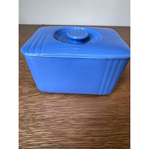 May include: A rectangular blue ceramic butter dish with a matching lid. The lid features a circular handle in the center and subtle vertical ribbed detailing on the corners. The base of the dish has a smooth finish with matching vertical ribs.