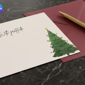 Puede incluir: Una tarjeta de felicitación con una ilustración de acuarela de un árbol de Navidad y la frase "With gratitude". Un sobre burdeos y un bolígrafo dorado están junto a la tarjeta. La tarjeta está sobre una superficie oscura.