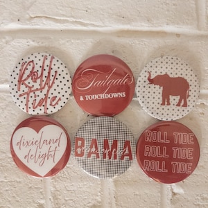Puede incluir: Seis botones redondos con varios diseños y texto. Los botones presentan frases como "Roll Tide", "Tailgates & Touchdowns" y "Dixieland Delight". La combinación de colores incluye rojo, blanco y negro.
