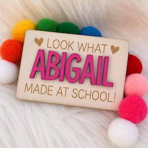 Pode incluir: Placa de madeira com o nome "ABIGAIL" em rosa, rodeada por bolas de feltro coloridas. A placa diz "LOOK WHAT ABIGAIL MADE AT SCHOOL!" em uma fonte divertida, com detalhes de coração. O fundo é uma superfície branca e fofa.