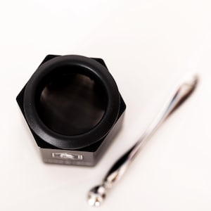 May include: A black hexagonal container with a circular opening and a fine mesh screen inside, accompanied by a slender, polished silver-toned metal spoon. The container features a small white logo on one of its flat side panels.