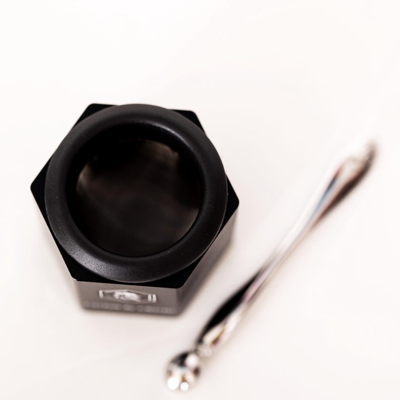 May include: A black hexagonal container with a circular opening and a fine mesh screen inside, accompanied by a slender, polished silver-toned metal spoon. The container features a small white logo on one of its flat side panels.