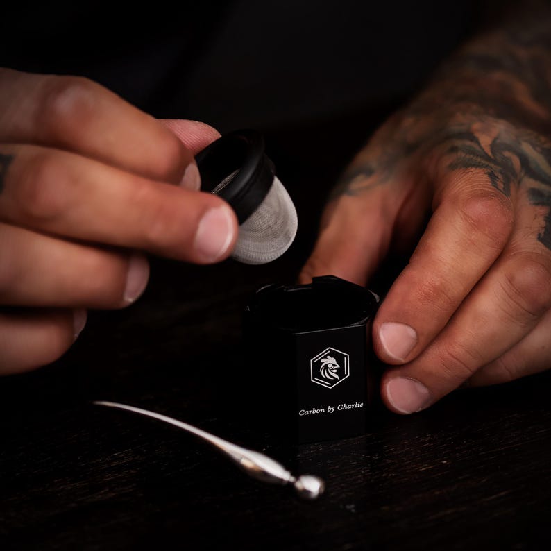 Person holding a mesh insert above a black Carbon by Charlie grinder with a small metal tool on a dark tabletop.