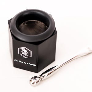 May include: A black hexagonal container with a mesh screen insert and a silver-colored metal tool featuring a spherical end. The container is printed with a white logo of a stylized bird head inside a hexagon and the text Carbon by Charlie.