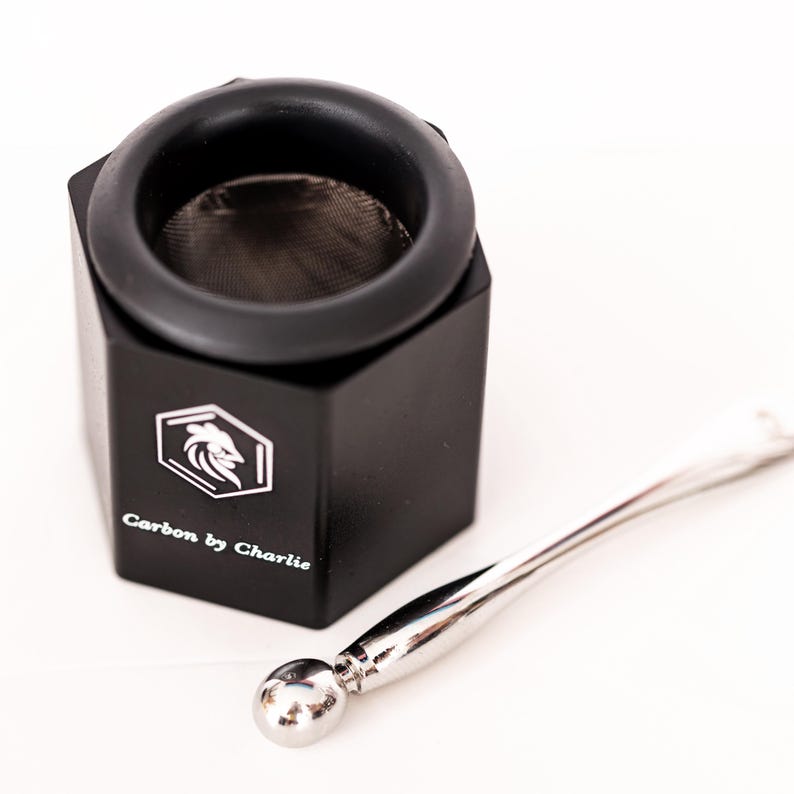 May include: A black hexagonal container with a mesh screen insert and a silver-colored metal tool featuring a spherical end. The container is printed with a white logo of a stylized bird head inside a hexagon and the text Carbon by Charlie.