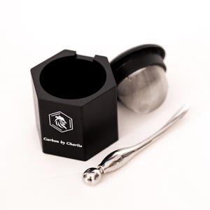 May include: A black hexagonal container with the text Carbon by Charlie and a rooster logo, a detached black lid with a silver mesh tea infuser, and a polished silver metal stirring tool with a spherical end piece.