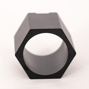 May include: A matte black metal component featuring a hexagonal exterior profile and a circular hollow center. The object has a uniform dark finish and a geometric shape with six flat sides, designed as a structural or mechanical part.