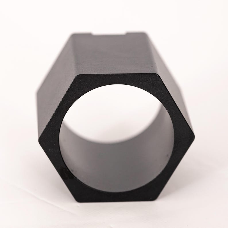May include: A matte black metal component featuring a hexagonal exterior profile and a circular hollow center. The object has a uniform dark finish and a geometric shape with six flat sides, designed as a structural or mechanical part.