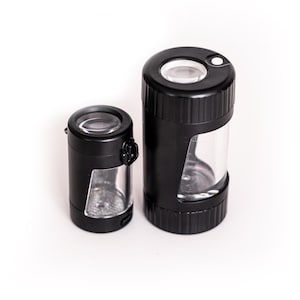May include: Two black magnifying glasses with clear cylindrical bodies. The larger one has a white button on top. Both have a lens at the top and a clear window to view the object being magnified. The magnifying glasses are on a white background.