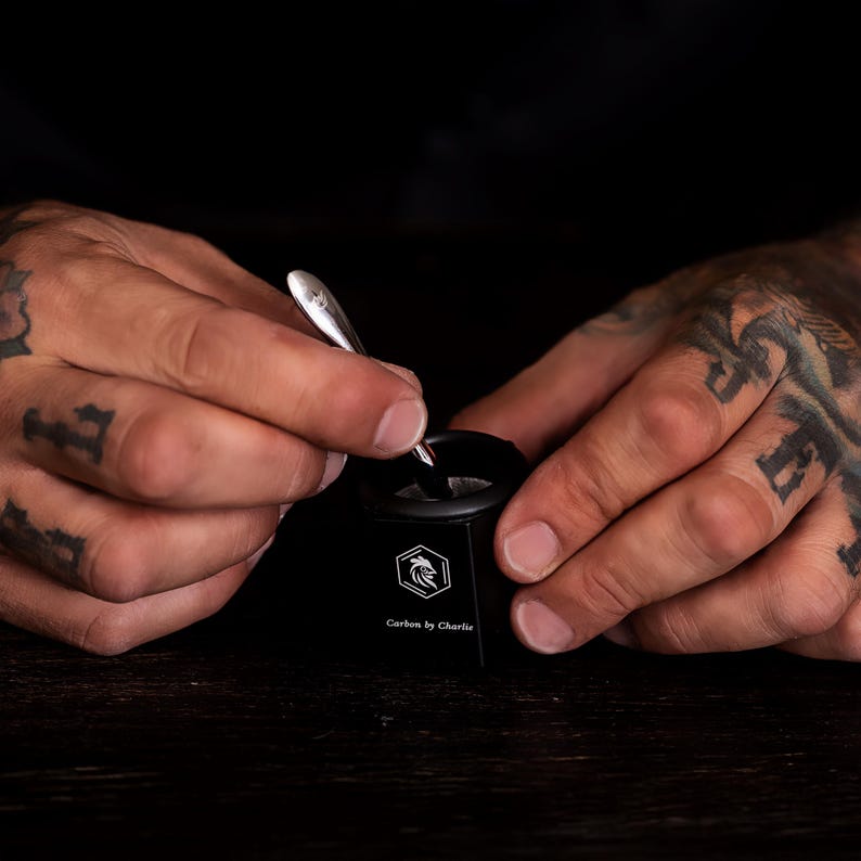 May include: A small black cylindrical container with a white hexagonal logo featuring a rooster head and the text Carbon by Charlie, paired with a slender silver metal spoon being used to stir the contents inside the container.