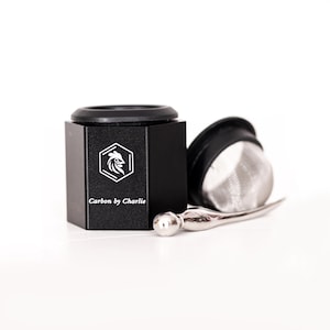 May include: A black, hexagonal container with a black lid and a silver metal tool. The container has a white logo and the text "Carbon by Charlie". A silver mesh filter is next to the container.