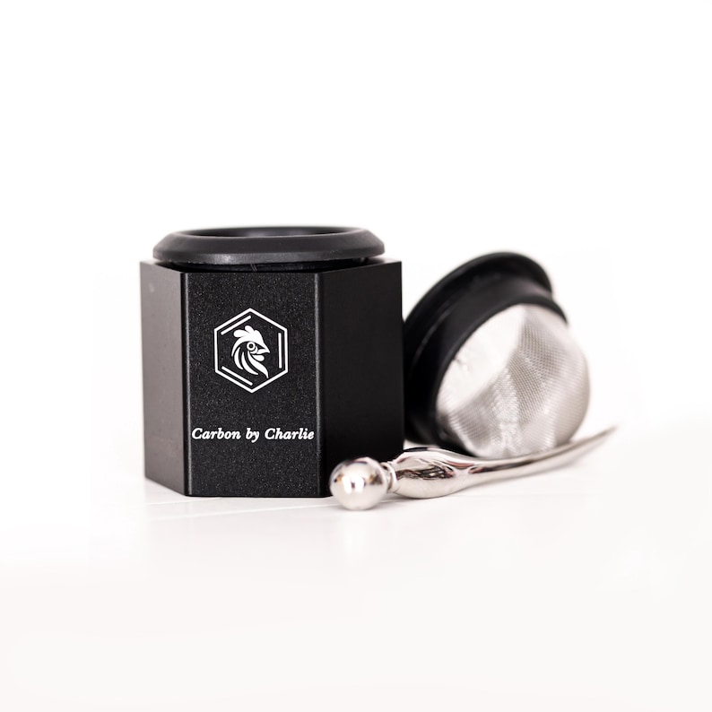 Black metal grinder with a mesh insert and small metal tool displayed on a white background, branded Carbon by Charlie.