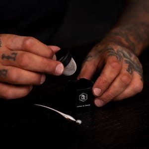May include: A black cylindrical container with a white logo featuring a rooster head and the text Carbon by Charlie. A small black lid with a silver mesh screen is held above the container, and a thin silver tool lies on the surface nearby.