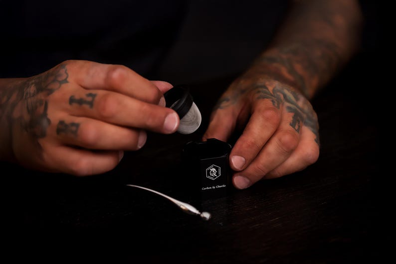 May include: A black cylindrical container with a white logo featuring a rooster head and the text Carbon by Charlie. A small black lid with a silver mesh screen is held above the container, and a thin silver tool lies on the surface nearby.