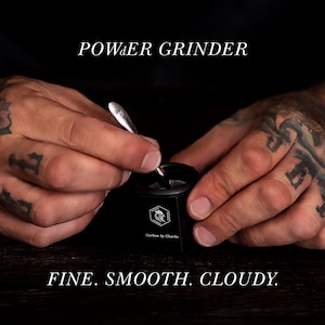 Hands using a small Carbon by Charlie grinder with a metal tool on a dark tabletop, with text overlay reading ‘POWDER GRINDER’ and ‘FINE. SMOOTH. CLOUDY.’