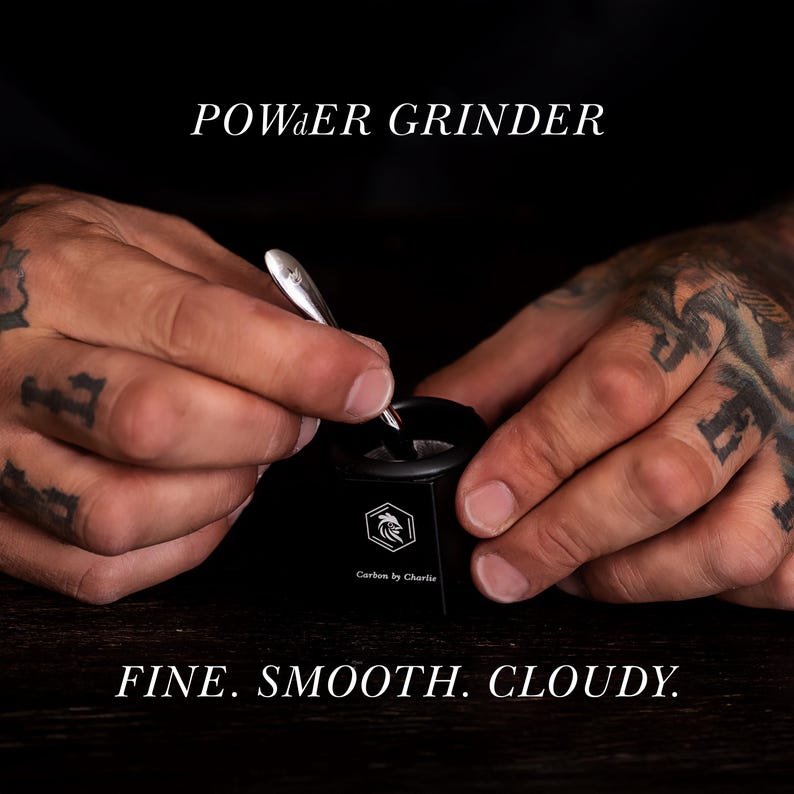 Hands using a small Carbon by Charlie grinder with a metal tool on a dark tabletop, with text overlay reading ‘POWDER GRINDER’ and ‘FINE. SMOOTH. CLOUDY.’