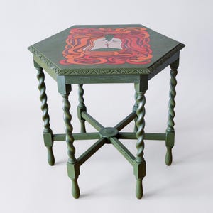 Op de afbeelding: Een groene, zeshoekige bijzettafel met een decoratief blad met het gezicht van een roodharige vrouw. De tafel heeft gesneden randen, gedraaide poten en is ongeveer 61 cm hoog.