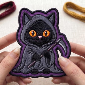 May include: An embroidered patch featuring a black cat wearing a grey hooded cloak and holding a scythe. The cat has large orange eyes and a small pink nose. The patch has a purple border and is surrounded by four small skeins of embroidery thread.