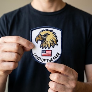May include: A white patch with a detailed golden eagle design, a red and white American flag, and the text "LAND OF THE FREE" in a navy blue border. The patch is held up against a black shirt.