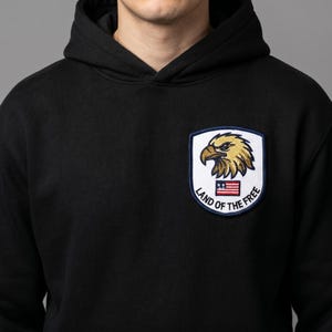 May include: Black hooded sweatshirt with a patch featuring a golden eagle, the American flag, and the text "Land of the Free". The patch is bordered in navy blue. The sweatshirt is made of a soft material.