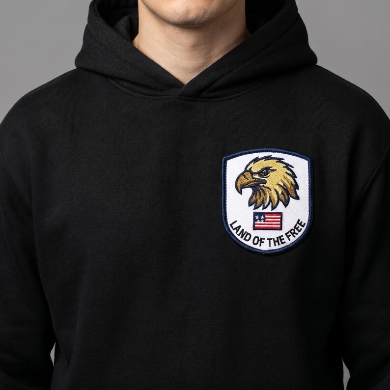 May include: Black hooded sweatshirt with a patch featuring a golden eagle, the American flag, and the text "Land of the Free". The patch is bordered in navy blue. The sweatshirt is made of a soft material.