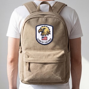May include: A tan backpack with a front pocket and adjustable straps. The backpack features a patch with a golden eagle, the American flag, and the text "Land of the Free."