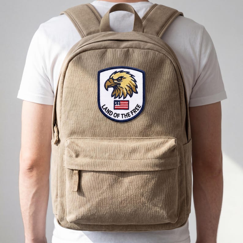 May include: A tan backpack with a front pocket and adjustable straps. The backpack features a patch with a golden eagle, the American flag, and the text "Land of the Free."