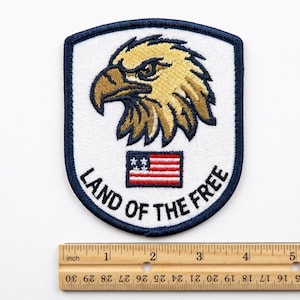 May include: Embroidered patch featuring a detailed eagle head in shades of gold and brown, set against a white background. The patch includes a small American flag and the text "LAND OF THE FREE" in black. The patch is bordered with a navy blue trim.