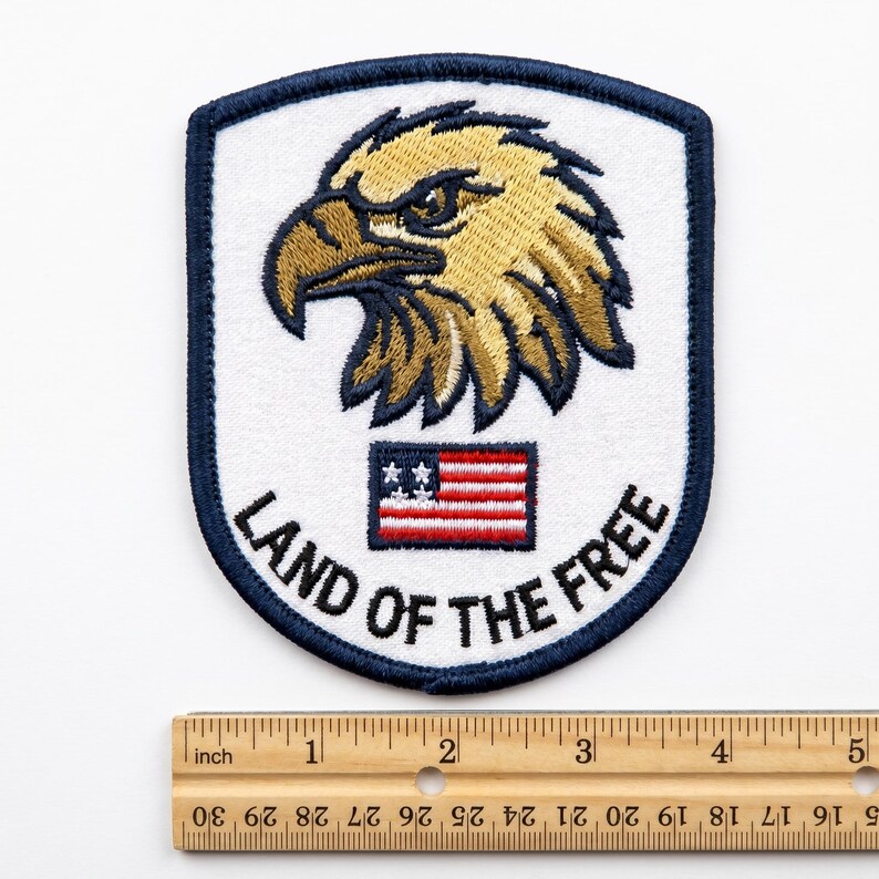 May include: Embroidered patch featuring a detailed eagle head in shades of gold and brown, set against a white background. The patch includes a small American flag and the text "LAND OF THE FREE" in black. The patch is bordered with a navy blue trim.