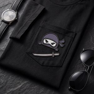 May include: A black t-shirt featuring a chest pocket with an embroidered purple ninja head and a silver sword. The shirt is displayed alongside a black wristwatch with a leather strap and a pair of black sunglasses with dark tinted lenses.