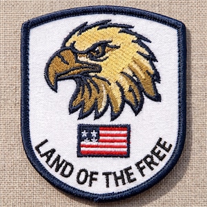 May include: Embroidered patch featuring a detailed golden eagle head above a stylized American flag. The patch is white with a navy blue border and the words "LAND OF THE FREE" in navy blue.